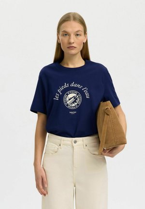 Vrouw met steil blond haar draagt een marineblauw T-shirt met print en beige broek met hoge taille, ze houdt een lichtbruine suède handtas onder haar arm.