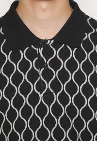 Polo noir avec un tissu texturé présentant un motif ondulé blanc, trois boutons au col et un design de col standard.