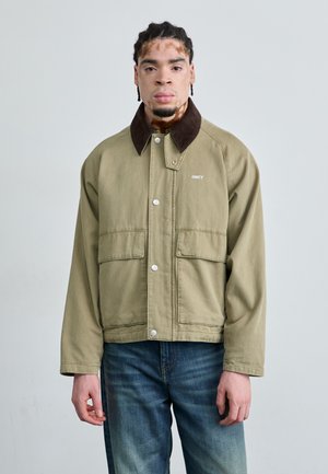 Olive green jacket with a brown corduroy collar, snap buttons, two front pockets, and the word "OBEY" embroidered in white. Worn with blue jeans.