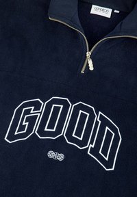 Marineblauer Fleece-Pullover mit einem Viertel-Reißverschluss-Kragen, versehen mit großen weißen "GOOD"-Buchstaben und einem runden Logo-Akzent. Weicher Stoff.
