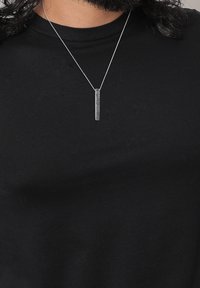 Pendentif argenté en forme de barre verticale avec un motif chevron sur une chaîne fine, porté sur un t-shirt noir à col rond par une personne aux cheveux foncés.