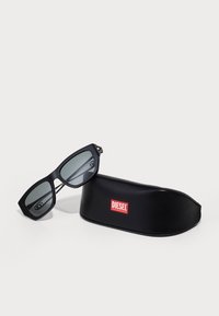 Óculos de sol retangulares pretos com lentes cinzas, apresentando um acabamento mate. Acompanhados por uma caixa rígida preta com o logótipo vermelho "DIESEL".