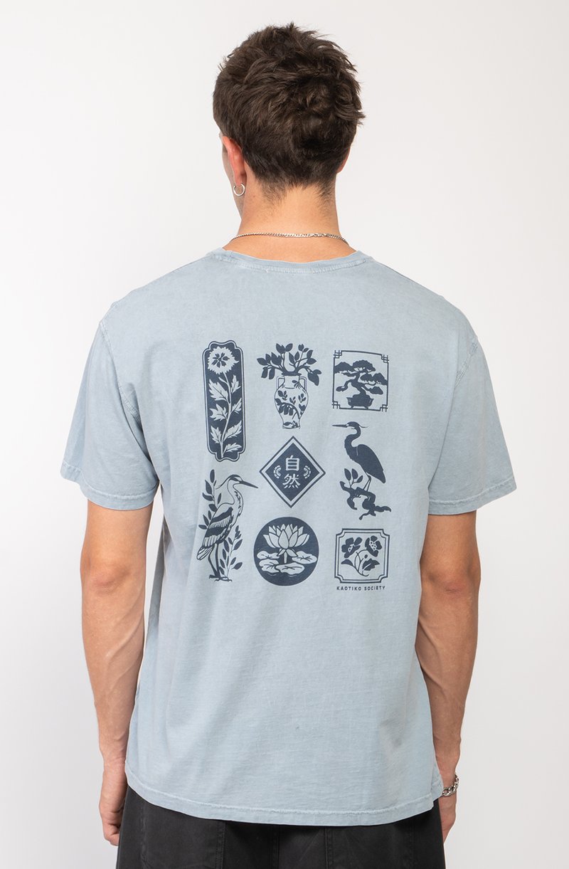 Camiseta de algodón de color azul claro con estampados gráficos oscuros de flores, pájaros y formas decorativas en la parte posterior. Mangas cortas y cuello redondo.