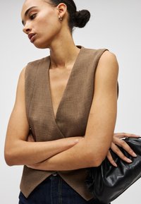 Γυναίκα με τα μαλλιά σε κότσο, φορώντας καφέ αμάνικο τοπ και σκούρο τζιν, κρατώντας μαύρο δερμάτινο τσαντάκι, με τα χέρια σταυρωμένα.