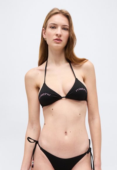 Jeune femme aux longs cheveux portant un bikini triangle noir avec le logo « misbhv » sur un fond uni gris clair.