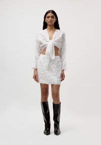 Crop top blanc texturé avec un détail à nouer à l'avant, associé à une mini-jupe blanche ajustée. Le mannequin porte des bottes noires à talons pointus jusqu'aux genoux.