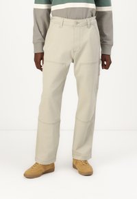 Beige cargo pants with large side pockets, visible stitching, and a relaxed fit, paired with light brown shoes.