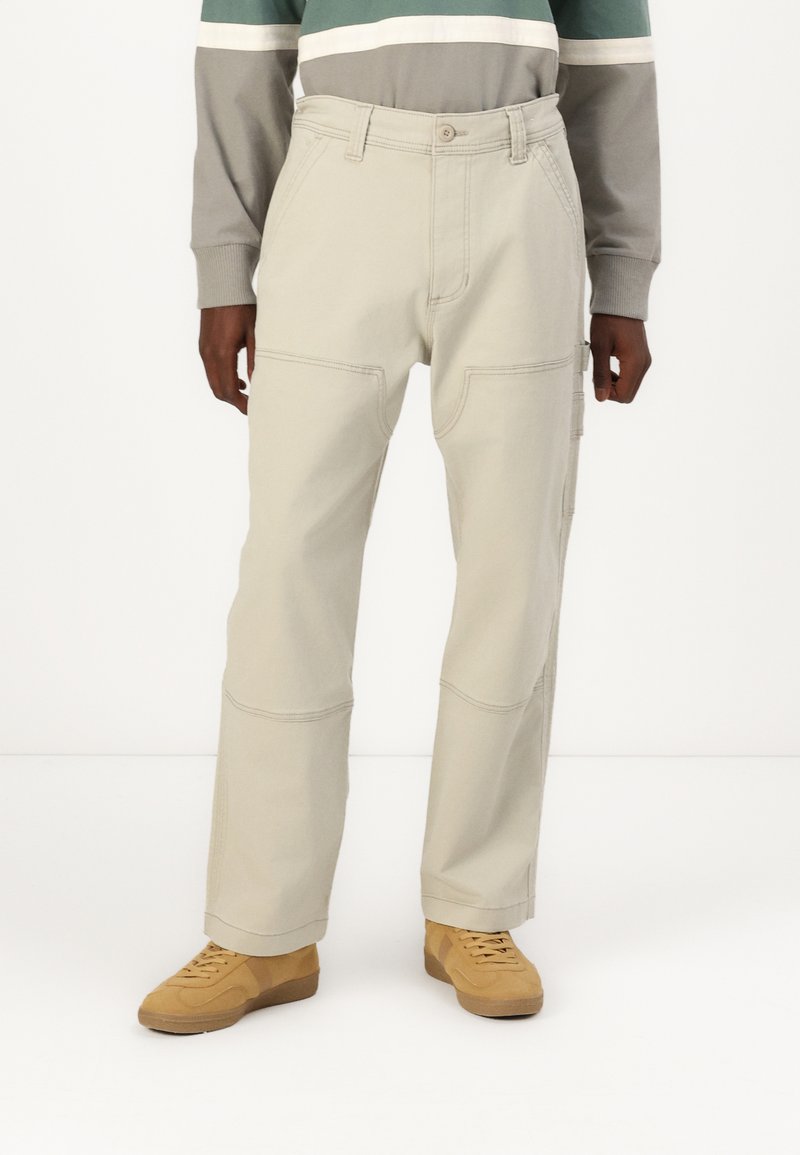 Beige cargo pants with large side pockets, visible stitching, and a relaxed fit, paired with light brown shoes.
