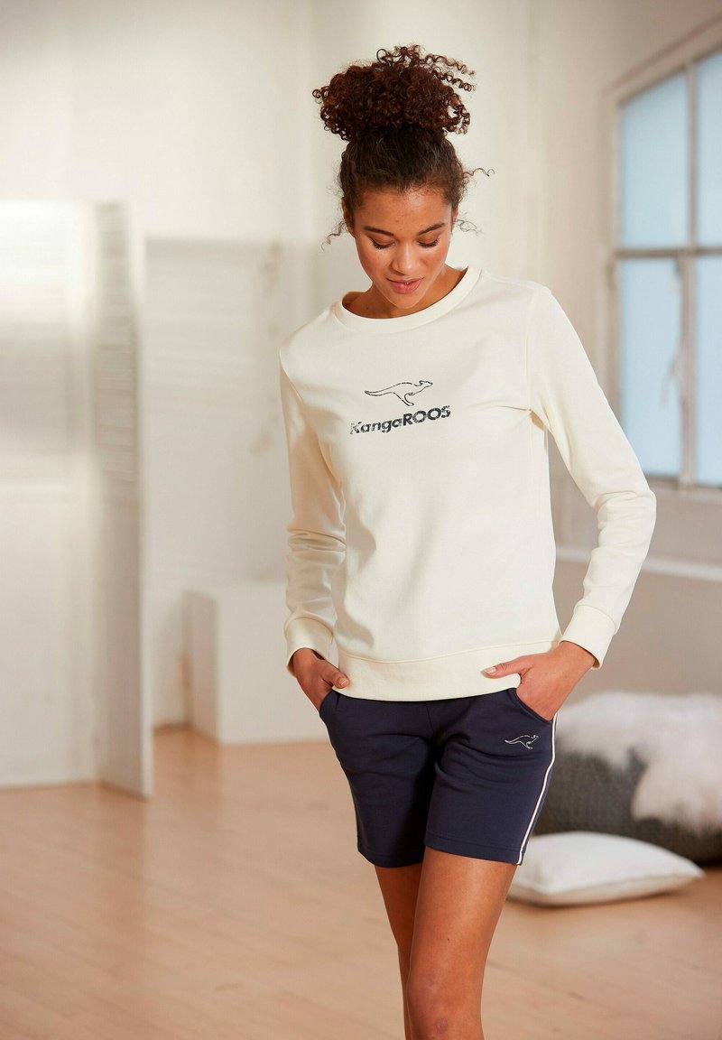Cremefarbenes Sweatshirt mit rundem Halsausschnitt und gesticktem Logo, kombiniert mit navyfarbenen Shorts mit weißen Seitenstreifen und Taschen.