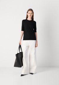 Svart kortermet topp kombinert med hvite vide bukser. Modellen holder en svart tote bag og står på en nøytral bakgrunn.