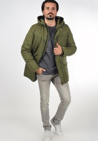 Chaqueta verde acolchada con capucha, frente con cremallera y botones a presión; llevada sobre un suéter gris y combinada con jeans gris claro y zapatillas blancas.