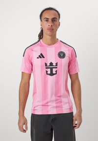 Rosa Sporttrikot mit vertikalen Streifen, ausgestattet mit einem schwarzen Wappen und Logo. Kurze Ärmel und schwarze Akzente an den Schultern. Leichtes Material.