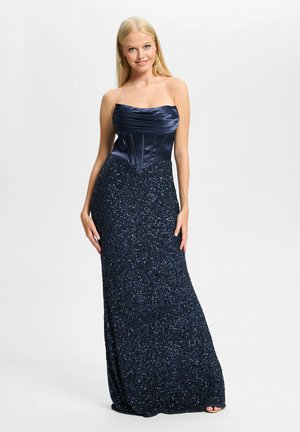 Frau mit langen blonden Haaren, die ein trägerloses dunkelblaues Satin- und Pailletten-Abendkleid trägt, lächelnd, vor schlicht weißem Hintergrund stehend.