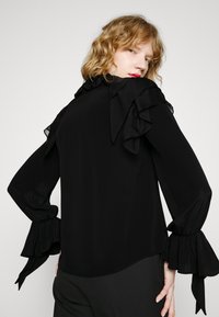 Blusa nera con colletto e polsini arruffati, maniche lunghe e un tessuto morbido e fluido con una texture minimale.