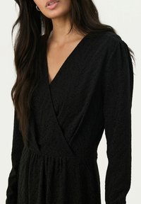 Schwarzes Kleid mit tiefem V-Ausschnitt, strukturiertem Stoff mit einem Blattmuster und langen Ärmeln, betont durch ein elastisches Taillendetail.