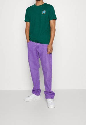 Maglietta verde a maniche corte con scollo rotondo e stampa grafica colorata, abbinata a jeans viola dal taglio ampio e sneakers bianche.