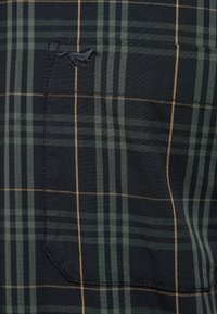 Dunkelgrüner und schwarzer Karostoff mit dünnen orangefarbenen Linien, ausgestattet mit einer gesteppten Tasche und einem kleinen schwarzen gestickten Pferdelogo in der oberen linken Ecke.