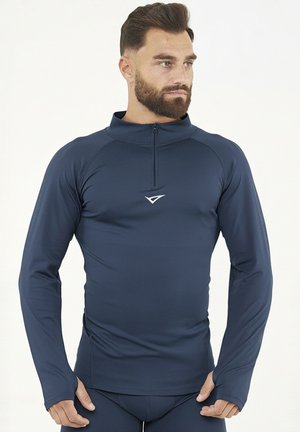 Bärtiger Mann trägt ein eng anliegendes navyblaues langärmeliges Sportoberteil mit einem Viertelreißverschluss und Daumenlöchern, steht vor einem einfarbigen Hintergrund.