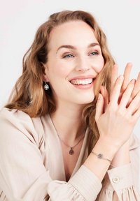 Zilveren boom des levens hanger ketting en bijpassende oorbellen, gedragen met een beige blouse. De armband heeft een vergelijkbare cirkelvormige charme. 
