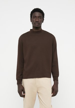 Bruine lange mouwen pullover met een hoge hals, gemaakt van zachte stof. Heeft een ontspannen pasvorm en geribde manchetten, gecombineerd met lichtbeige broeken.