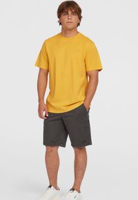 Gul bomulds T-shirt med korte ærmer, parret med grå shorts. Skoene er hvide med sorte accenter. Enkel og casual design.
