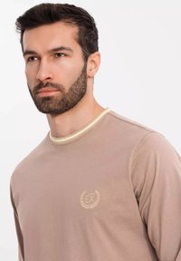 Beige pitkähihaisessa paidassa on pyöreä kaula-aukko ja vaaleat kontrastit. Rinnassa on pieni brodeerattu logo. Pehmeä ja sileä rakenne.