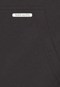 Schwarzer Stoff mit einer Tasche und einem kleinen metallic beschrifteten Etikett "bazix republiq". Die Textur wirkt weich und glatt.