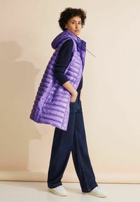 Lila vadderad väst med huva, som har horisontell quiltning, framlommor och en stilren design. Matchad med mörk denim flares och vita sneakers.