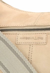 Sac en daim beige avec fermeture éclair, surface texturée, sangle en tissu gris et étiquette portant la mention "HARBOUR 2ND" avec un emblème d'ancre.