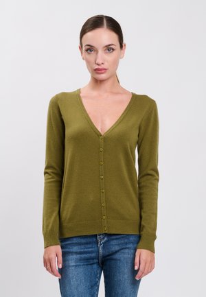 Kvinde med lys hud og håret bundet tilbage iført en olivengrøn cardigan med knapper og blå jeans, der står mod en ensartet lys baggrund.