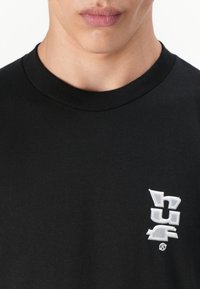 Sort bomulds T-shirt med rund halsudskæring og et lille hvidt broderet logo på venstre bryst. Glat tekstur, afslappet design.