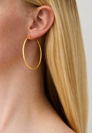 Nahaufnahme eines Frauenohrs mit glattem, blondem Haar, das einen großen, dünnen goldenen Creolenohrring trägt.