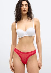 Γυναίκα με καστανά σγουρά μαλλιά, φοράει λευκό στράπλες σουτιέν και κόκκινο δαντελωτό εσώρουχο με μικρά καρδιακά σχέδια, στέκεται μπροστά σε ένα απλό φόντο.