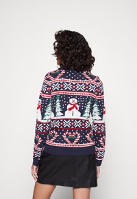 Femme portant un pull de vacances bleu marine avec des bonhommes de neige, des sapins de Noël, des cannes en sucre d'orge et des motifs de flocons de neige, assorti à une jupe noire.