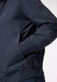 Giacca bomber blu navy con tasca con zip sulla manica, polsini elasticizzati, tasche laterali e etichetta testurizzata sul fondo. 