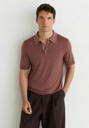 Man with short dark hair wearing a brown short-sleeve collared shirt and dark brown pants, standing against a white wall.