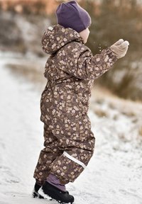 Combinaison de neige marron à motif floral avec un design matelassé, accompagnée d'un bonnet violet et de moufles beige, debout sur un sol enneigé.