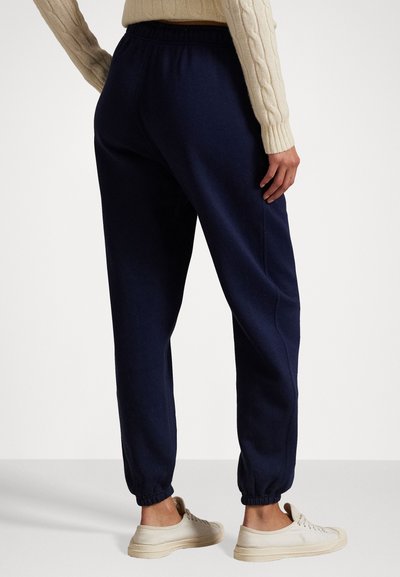 Navy sweatpants with elastic waistband and cuffs, featuring a soft texture. Worn with light-colored sneakers, shown from behind.