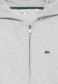 Siva zip-up dukserica od mekanog materijala, s kapuljačom obloženom zelenom tkaninom i izvezenom krokodilskom logotipom na lijevoj strani grudnog koša. Vidljivi su detalji za zatvaranje na patentnom zatvaraču.