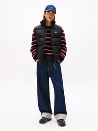 Svart pufferväst över en randig tröja i röd, marinblå och vit. Vida mörka denimjeans med kontrasterande fållar och svarta sneakers.