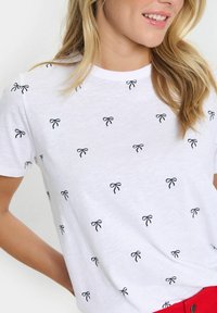 Balts kokvilnas T-krekls ar teksturētu apdari, kurā ir izkaisītas melnas izmēģinājuma lentes. Īsas piedurknes un klasiskā apaļā kakla dizains.