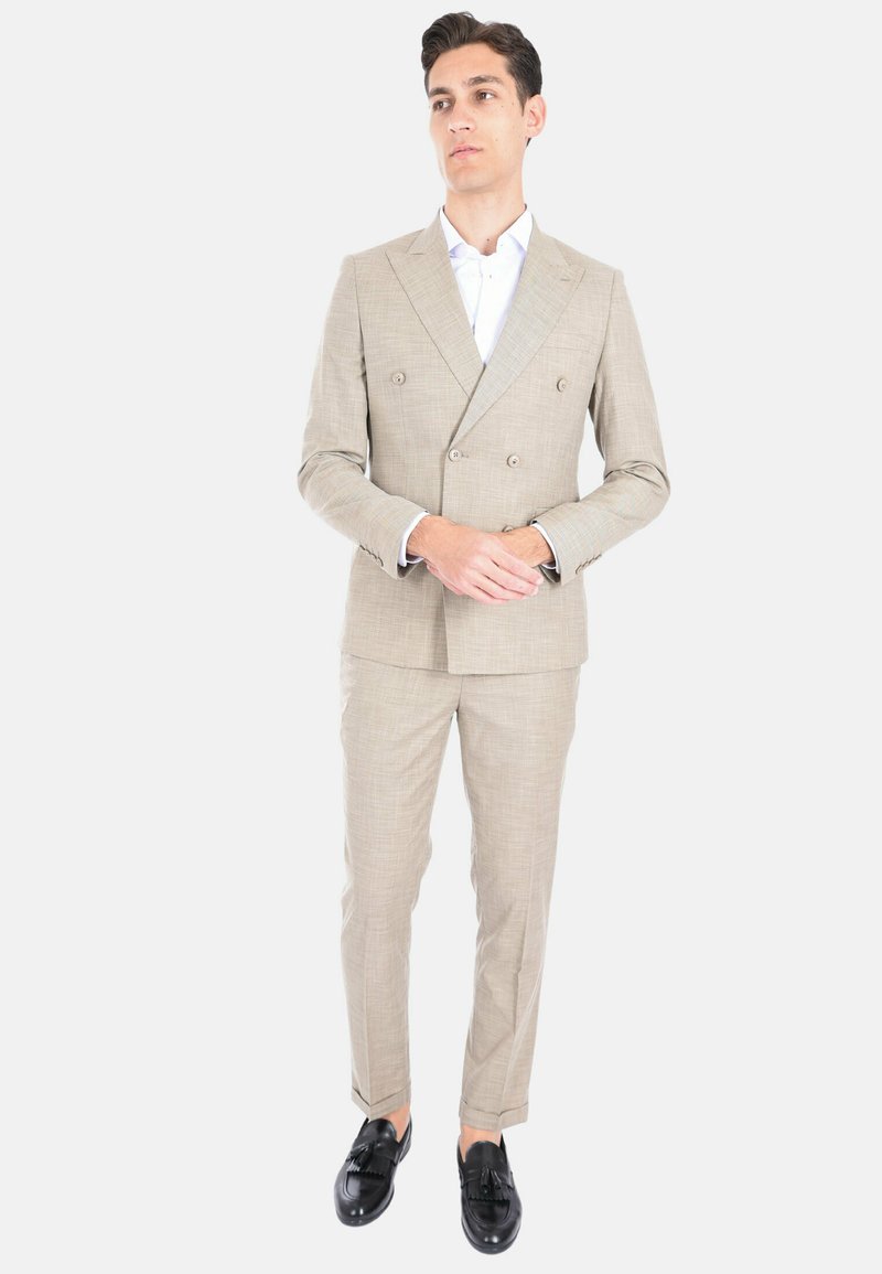 Completo doppiopetto beige chiaro con tessuto testurizzato, taglio aderente, abbinato a una camicia bianca fresca e mocassini neri.