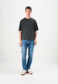 Young man standing, wearing a loose dark gray t-shirt, light blue jeans, white sneakers, with a tattoo visible on his left forearm.