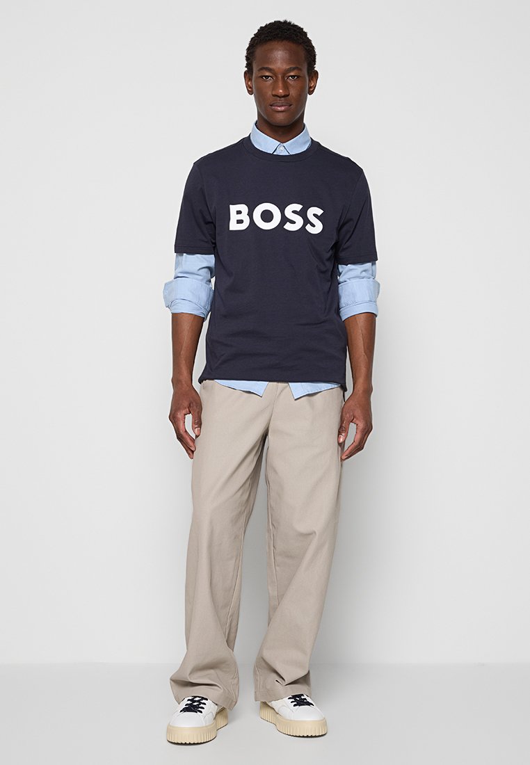 T-shirt bleu marine avec un grand texte blanc "BOSS", superposé sur une chemise boutonnée bleu clair, associé à un pantalon beige et des baskets blanches.