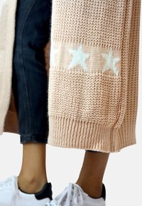 Cárdigan de punto en rosa claro con dobladillo acanalado y patrón de estrellas. Combinado con jeans oscuros y zapatillas blancas, que muestra un look casual.