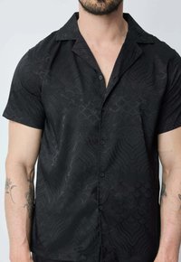 Chemise noire à manches courtes avec un motif ondulé subtil, tissu léger, fermeture à boutons sur le devant et col classique.