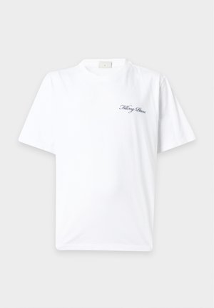 Jeune homme portant un t-shirt blanc avec texte, pantalon gris ample et délavé, et baskets noires et blanches, debout avec une main dans la poche.