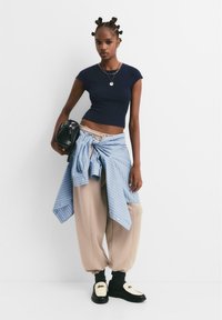 Námornícky priliehavý crop top, béžové tepláky s manžetami, modré pruhované flanelové košele viazané okolo pása, čierna kabelka a čierne topánky s krémovými akcentmi.