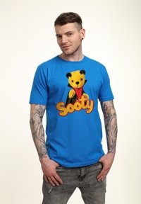 Modré bavlněné tričko s grafikou žlutého medvěda s černýma ušima a červeným motýlkem, doplněné textem "Sooty" v červené a žluté barvě.