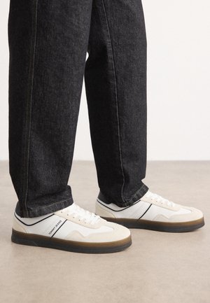 Białe trampki z czarną podeszwą i logo "Tommy Jeans" z boku, noszone z czarnymi jeansami, stojące na jasnej podłodze.